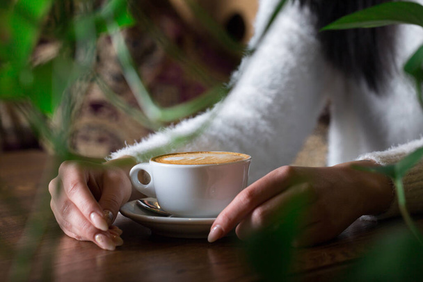 Egy lány ül az asztalnál egy csésze forró cappuccinóval a kezében. - Fotó, kép
