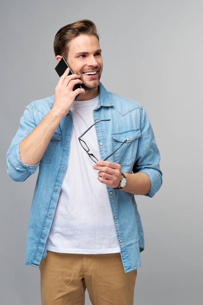pretty casual man in blue jeans shirt holding his phone standing over studio grey background - Foto, afbeelding