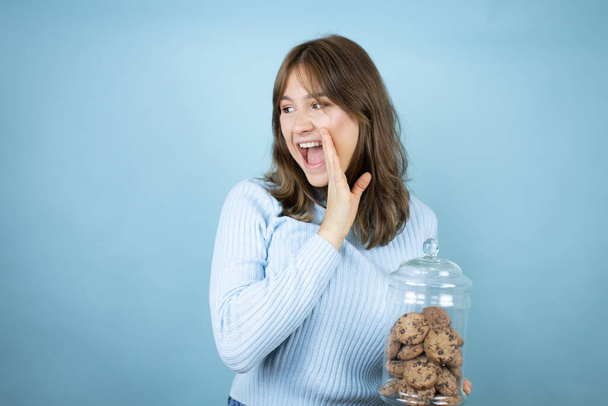 Jovem mulher bonita segurando chocolate chips cookies jar sobre isolado fundo azul mão na boca dizendo rumor secreto, sussurrando conversa maliciosa - Foto, Imagem
