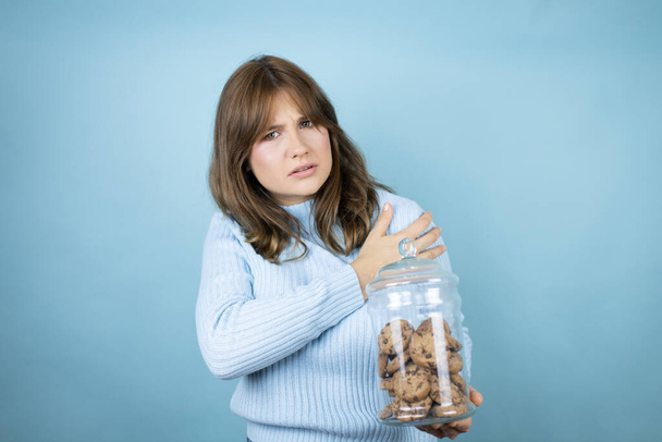 Молода красива жінка тримає шоколадні чіпси банку печива над ізольованим синім тлом з болем на її плечі і болючим виразом
 - Фото, зображення
