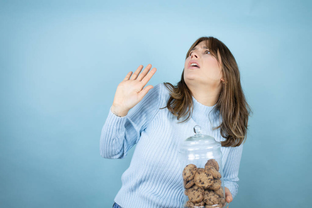Jonge mooie vrouw met chocolade chips koeken pot over geïsoleerde blauwe achtergrond bang met haar armen omhoog als iets vallen van boven - Foto, afbeelding