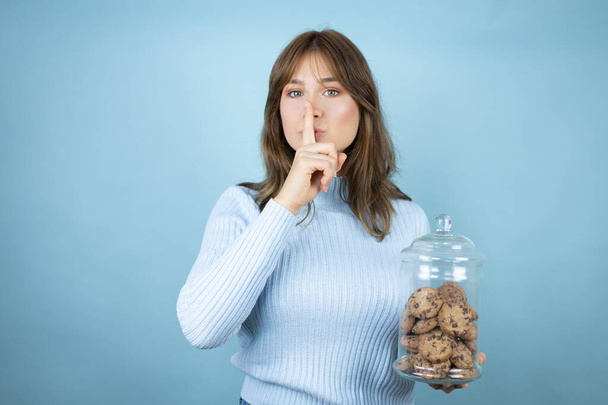 Молодая красивая женщина, держащая банку шоколадных чипсов на изолированном синем фоне, просит тишины с пальцем на губах. Молчание и тайная концепция. - Фото, изображение