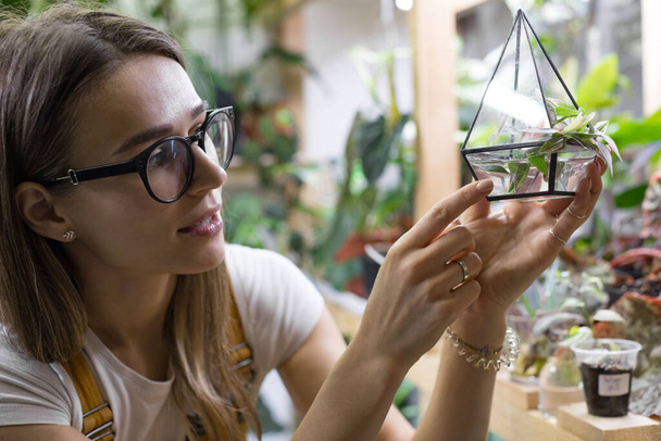 Fiatal nő kertész szemüvegben nézi dugványok tradescantia egy kis üveg florarium a növény csírázás, cserepes szobanövények a háttérben.Otthon kertészkedés, szabadúszó, hobbi  - Fotó, kép