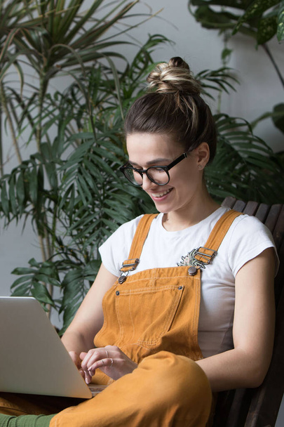 Young female gardener in glasses wearing overalls, sitting on wooden chair in greenhouse, using laptop after work, communicates on Internet with customer. Plant on background.Home gardening, freelance - Photo, Image