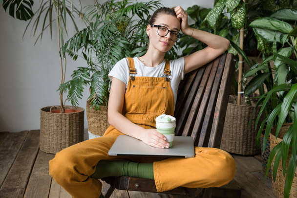 Mujer jardinero europeo vistiendo overoles, descansando después del trabajo, sentado en la silla de madera en el invernadero de casa, mantenga taza de café / té reutilizable, mirando a la cámara con el ordenador portátil en sus rodillas. Descanso durante el trabajo - Foto, imagen