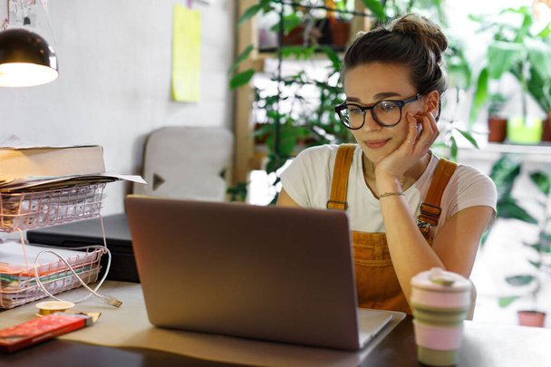 Young female gardener in glasses using laptop, communicates on internet with customer in home garden/greenhouse, reusable coffee/tea mug on table.Cozy office workplace, remote work, E learning concept - Photo, Image