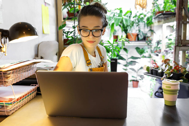 Młoda kobieta ogrodnik w okularach za pomocą laptopa, komunikuje się w Internecie z klientem w ogrodzie / szklarni, kubek do kawy wielokrotnego użytku na stole.Przytulne miejsce pracy biurowej, praca zdalna, Koncepcja e-learningu - Zdjęcie, obraz
