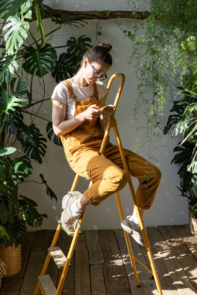 Gärtnerin in orangefarbenem Overall sitzt auf einer Leiter in ihrem Gewächshaus, ruht sich aus, benutzt Smartphone, liest Nachrichten am Telefon. Pause während der Arbeit - Foto, Bild