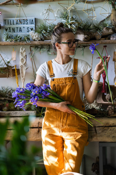 Überraschte Floristin mit Brille, trägt orangefarbene Overalls, umgeben von einer Ansammlung von Tillandsia-Pflanzen, bezaubert mit angenehmen Geschenken, lächelt und blickt auf einen Strauß Iris-Blumen. Indoor-Garten - Foto, Bild