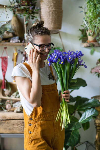 Mujer florista recibe saludos de cumpleaños de amigos y clientes por teléfono en su trabajo rodeado de plantas de interior, encantado con un regalo agradable, sonriendo, la celebración de ramo de flores de iris. - Foto, imagen