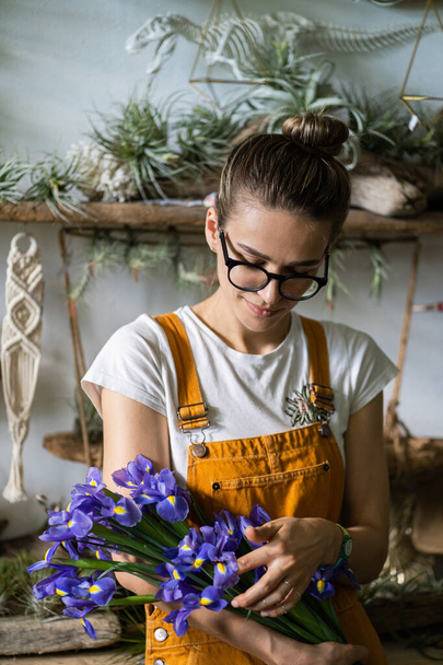 Verrast bloemist vrouw in bril, draag oranje overalls omgeven door collectie van tillandsia luchtplanten, gecharmeerd met aangename gift, glimlachen, kijken naar een boeket irissen bloemen. Tuin binnen - Foto, afbeelding