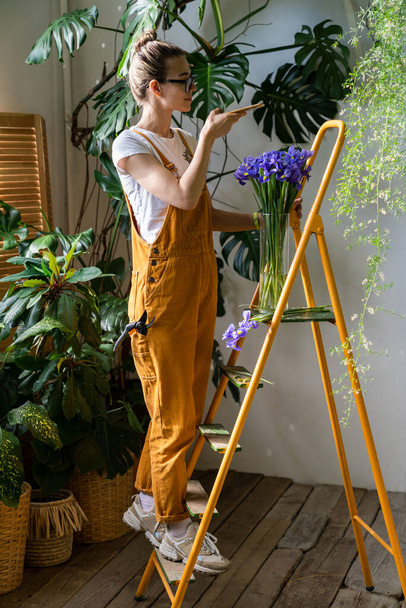 Smiling woman florist in orange overalls standing on stepladder, takes a photo of bouquet of purple irises in glass vase on smartphone. Love for flowers, hobby, indoor gardening - Photo, Image