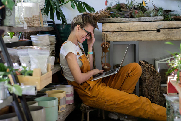 Jardineiro mulher bonito em óculos usar macacão laranja, sentado no chão na loja de flores usando laptop após o trabalho, sorrindo e falando em videochamada cercada por plantas. Casa jardinagem, freelance - Foto, Imagem