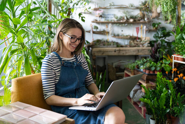 Vrouwelijke tuinman met bril draagt blauwe jeans overalls, zit op een stoel in de kas, gebruikt laptop na het werk, communiceert op internet met klanten omringd door planten. Huis tuinieren, freelance. - Foto, afbeelding