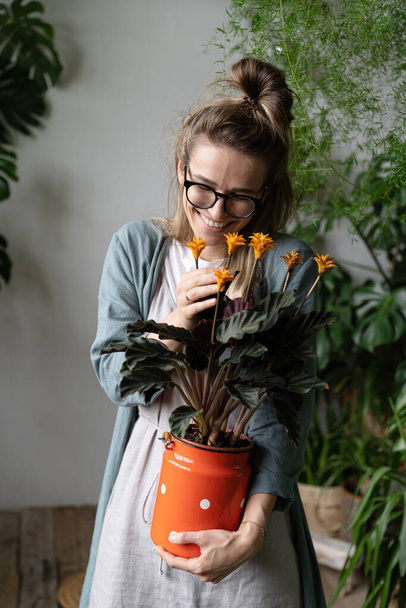 Young smiling woman gardener in eyeglasses wearing linen dress, holding a flowering calathea plant in old red milk can standing in her home garden. Love of houseplants - 写真・画像