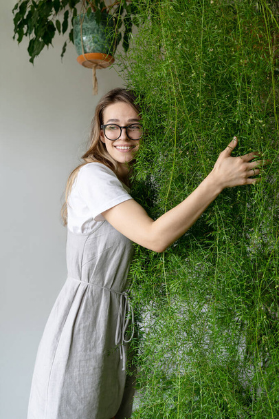 Ευτυχισμένη νεαρή γυναίκα κηπουρός με λιτό φόρεμα, αγκαλιάζει πλούσια σπαράγγια φτέρη στο ανθοπωλείο της. Πράσινο στο σπίτι. Η αγάπη για τα φυτά. Εσωτερικό άνετο κήπο. - Φωτογραφία, εικόνα
