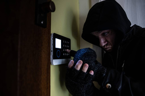 Een inbreker met handschoenen en een kap op zijn hoofd probeert het alarmsysteem uit te schakelen.. - Foto, afbeelding