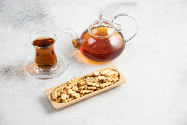 A glass teapot with a cup of tea and a wooden board full of crackers - Фото, изображение