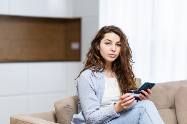 Portrait happy female shopper using instant easy mobile payments making purchase in online store young woman customer holding credit card and smartphone sitting on couch at home - Valokuva, kuva
