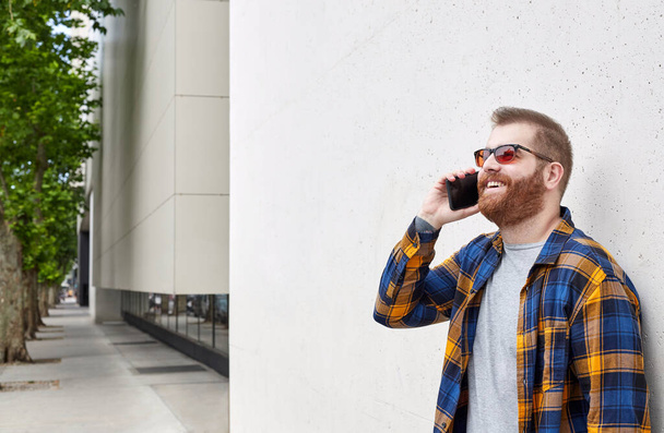 Joven hombre latino barbudo en el teléfono vestido con camisa de leñador de pie junto a la pared de hormigón al aire libre. Copiar espacio - Foto, imagen