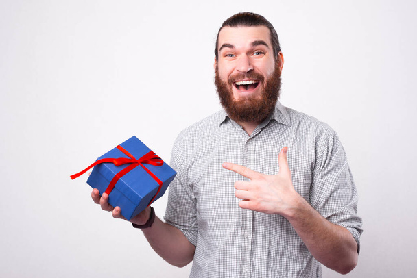 Очень взволнованный мужчина держит подарок, указывая на него и глядя в камеру - Фото, изображение