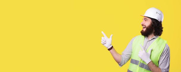 Um engenheiro barbudo animado está olhando sorrindo e apontando para um espaço livre perto de uma parede amarela - Foto, Imagem