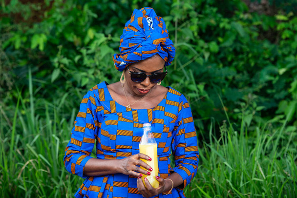 a beautiful mature african woman in traditional dress standing in the countryside looking at a bottle of fruit juice. - Photo, Image