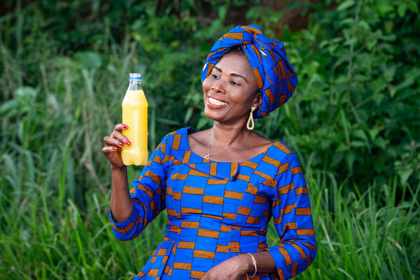 una hermosa mujer africana madura con ropa tradicional de pie en el campo mirando una botella de jugo de frutas sonriendo. - Foto, Imagen