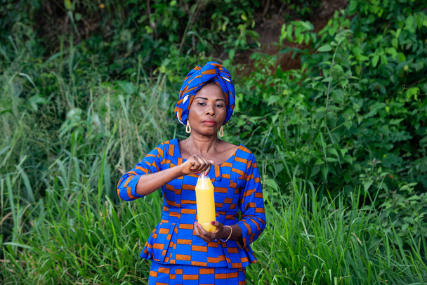 Geleneksel elbiseli, güzel, olgun bir Afrikalı kadın elinde meyve suyu şişesiyle kırsalda dikiliyor. - Fotoğraf, Görsel
