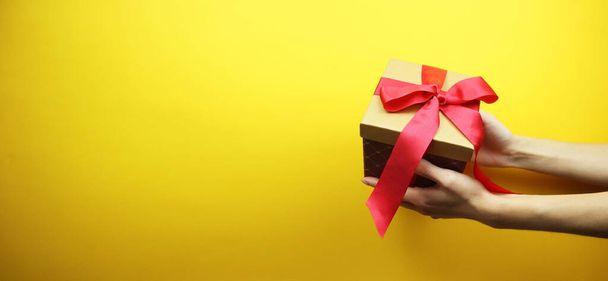 Cardboard box with a red ribbon and a bow in hands. Isolated on a yellow background. Place for your text. Copy space. Banner. - Photo, Image