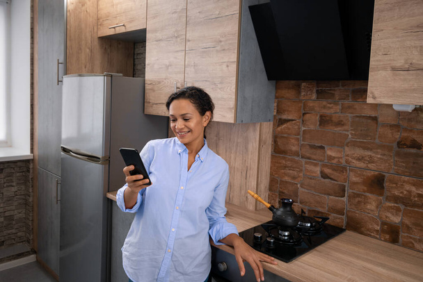 La jeune femme lit des SMS dans un smartphone alors qu'elle se tient debout dans la cuisine. - Photo, image