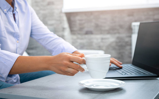 La vista de la mano de una mujer levantando una taza con un portátil en el fondo. laptop. Trabajo remoto desde casa. - Foto, Imagen