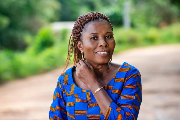 een mooie Afrikaanse vrouw in traditionele jurk die op het platteland staat en haar oorbel laat zien terwijl ze lacht. - Foto, afbeelding
