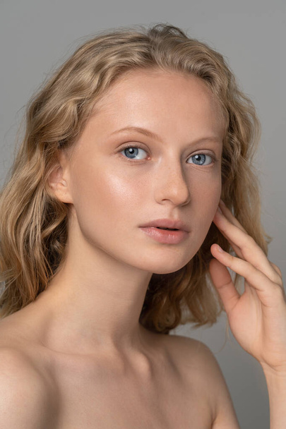 Closeup of pretty young woman face with blue eyes, curly natural blonde hair, has no makeup, touching her soft skin, standing shirtless with bare shoulders, looking at camera. Studio grey background - Foto, afbeelding