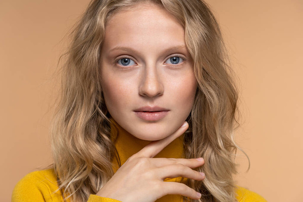Young woman face with blue eyes, curly natural blonde hair and eyebrows, has no makeup, looking at camera, touching chin. Girl with perfect fresh clean skin over studio beige background. Youth. - Photo, Image