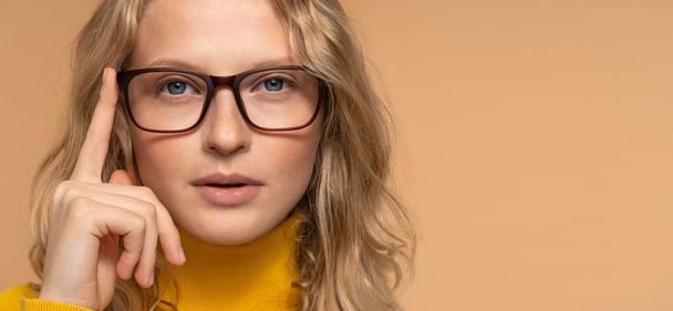 Optical store advertising, eyesight check. Close up portrait of young blonde woman in glasses looking at camera isolated over beige studio background, copy space. - 写真・画像