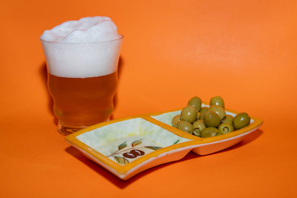 Пиво с большим количеством пены и оливок на оранжевом фоне - Фото, изображение
