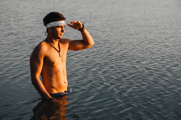 L'uomo con una muscolatura perfetta sta in acqua al tramonto. Luogo per testo o pubblicità. - Foto, immagini