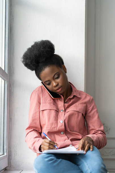 Fókuszált afro-amerikai biracial nő jegyzetel, tisztázza az információt, telefonon beszél, az ablakpárkányon ül. Biracial női munkavállalói tanácsadás mobil eszköz, dolgozik otthon távolról - Fotó, kép
