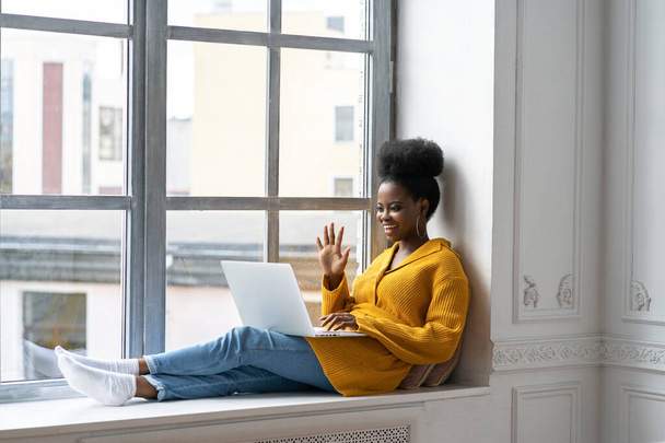 Uśmiechnięta Afroamerykańska milenijna kobieta z afro fryzurą w żółtym kardiganie siedzi na parapecie, odpoczywa, patrzy na kamerę internetową, mówi cześć, rozmawia na video czacie z przyjaciółmi na laptopie. - Zdjęcie, obraz