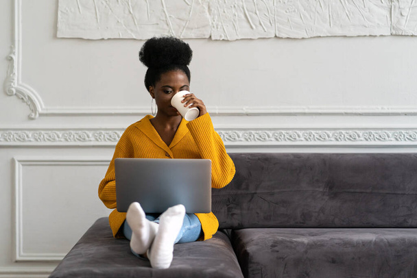Afro-American biracial woman with afro hairstyle in yellow cardigan sitting on couch, working online on laptop, watching webinar or videos in social media, holding mug, enjoying drinking coffee - Photo, Image