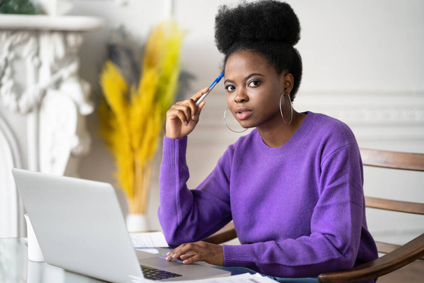 Portrait of Afro-American millennial student woman with afro hairstyle looking at camera while making researches browsing information on laptop, preparing for exam online, holding a pen. - Фото, зображення