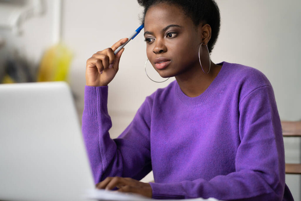 Afro-American millennial student woman with afro hairstyle browsing information on laptop, preparing for exam online, watching video course, holding a pen. Online education in zoom app, video training - Фото, зображення
