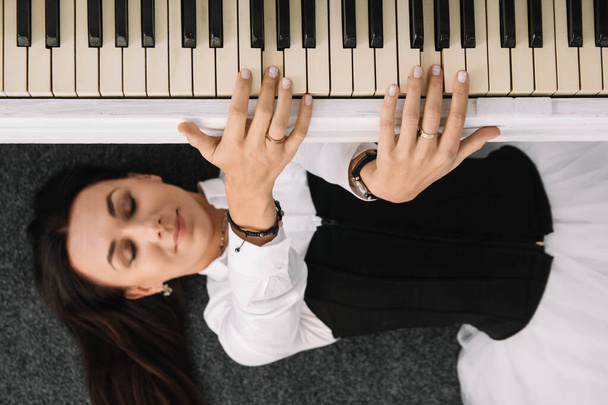 Beautiful woman dressed in a white dress with a black corset lies on the floor near white piano playing on the keys. Place for text or advertising. View from above. - Photo, Image