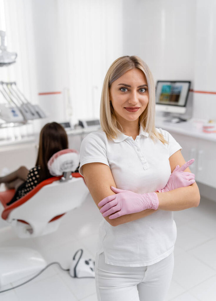 Blonde dentist doctor stands on patient in dentist chair background. Doctor in white scrubs at working place. - Фото, зображення