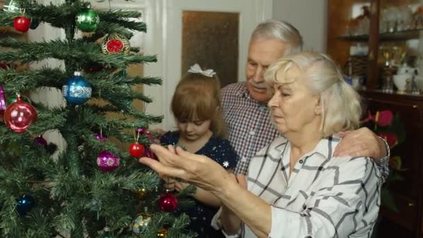 Mädchen mit Senioren-Oma, Opa schmückt künstlichen Weihnachtsbaum mit Ornamenten und Spielzeug - Filmmaterial, Video