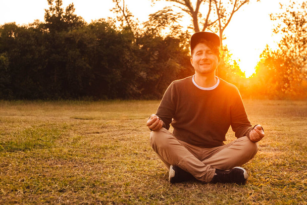 瞑想的な立場の若いハンサムな男は、森と日没の背景に芝生の上に座っています。自由のリラクゼーションの概念。テキストまたは広告の場所. - 写真・画像