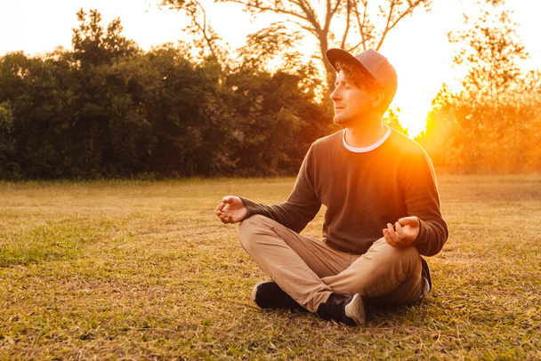 Jonge knappe man in een meditatieve positie zit op een grasveld op een achtergrond van bos en zonsondergang. Concept van vrijheid ontspanning. Plaats voor tekst of reclame. - Foto, afbeelding