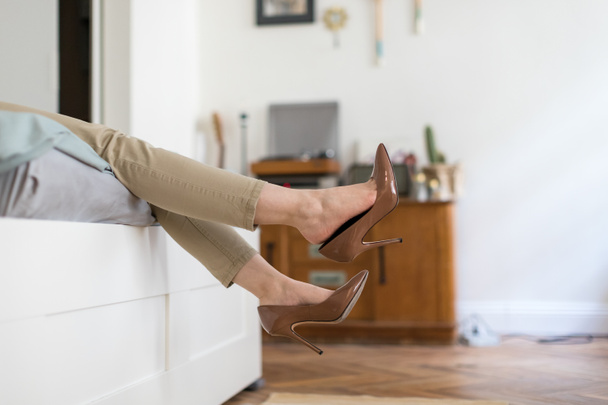 Η κουρασμένη γυναίκα ξεκουράζεται βγάζοντας τα καφέ ψηλοτάκουνα παπούτσια της μετά τη δουλειά ή περπατώντας, ξαπλωμένη στον καναπέ. άβολα παπούτσια - Φωτογραφία, εικόνα