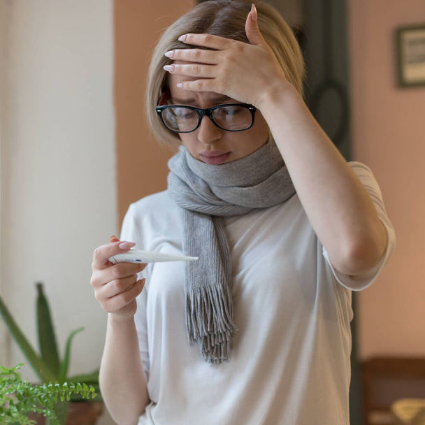 молода жінка в окулярах і сорочці, загорнута в шарф, має холодні симптоми, дивиться на термометр, торкається її чола. Вірус, сезон грипу
 - Фото, зображення